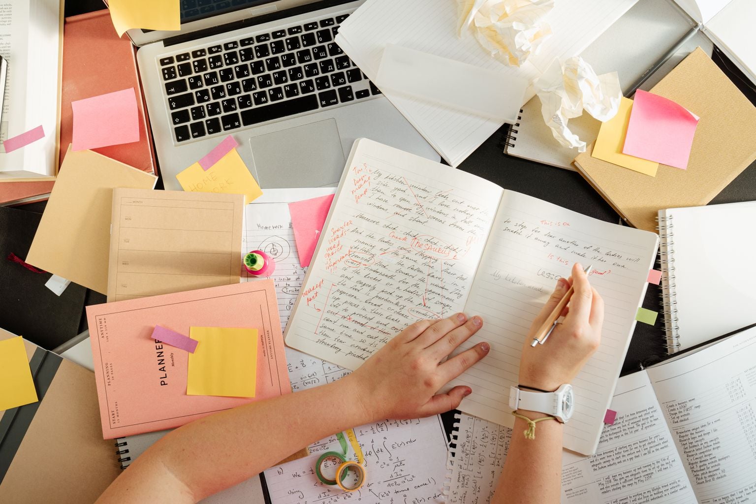 Overhead shot of a person organizing notes and study materials on a cluttered desk with a laptop