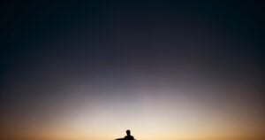 A lone figure sits on a bench at dusk, looking out over calm water toward a warm horizon.