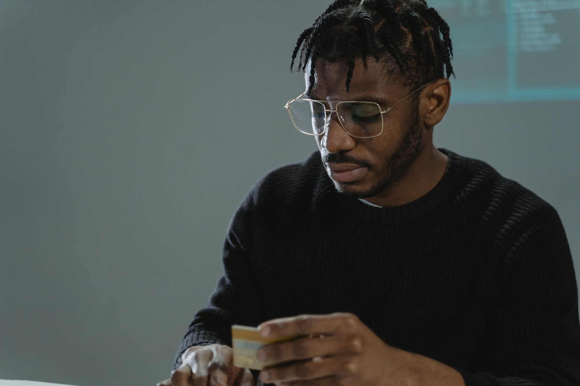 Focused man with eyeglasses holding a credit card in a dimly lit room, wearing a black sweater.