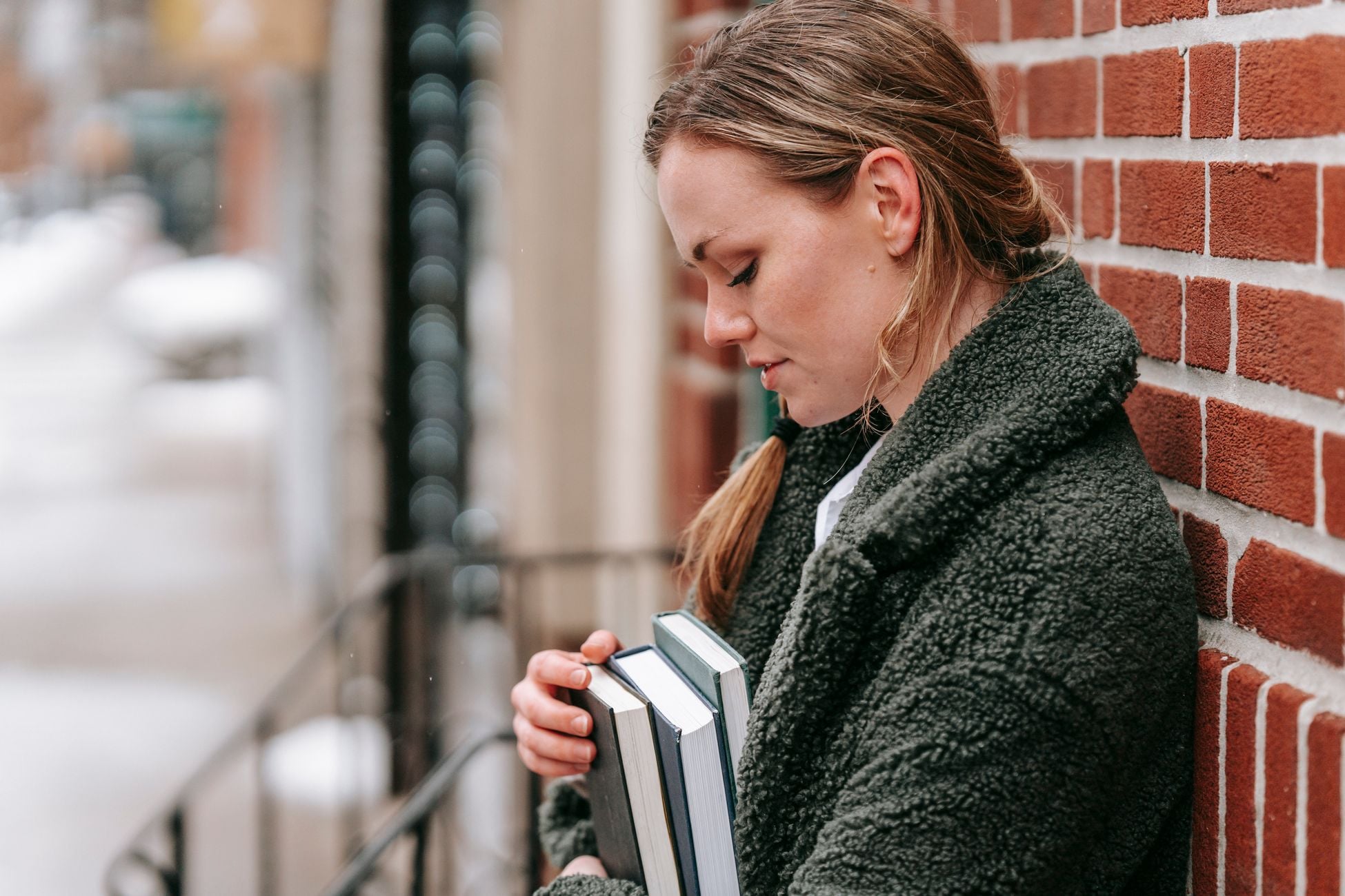 Side view of young female in outerwear with textbooks looking down against rough wall in town