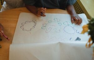 Child drawing with crayons at a table, with paper and art supplies visible.