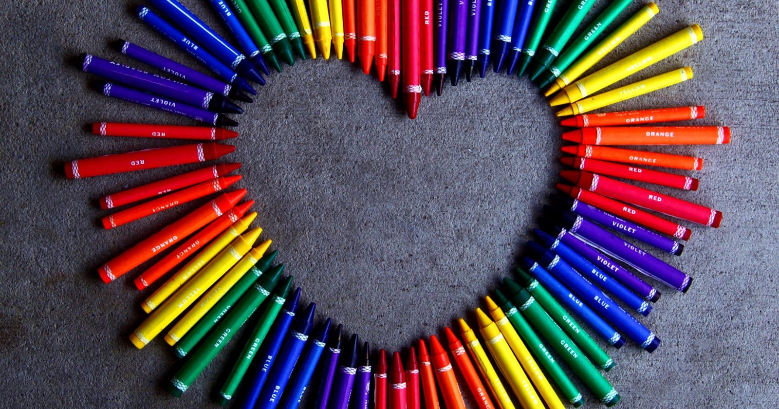 Colouring crayons in rainbow colours arranged in the shape of a heart on a grey surface
