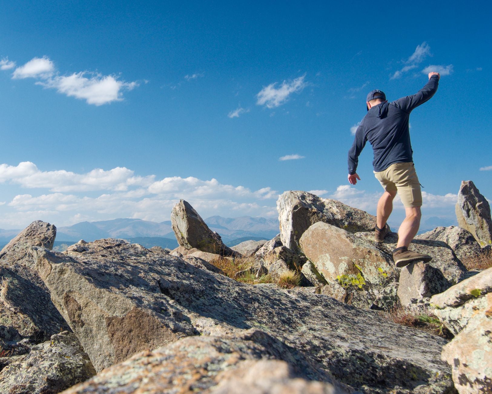 A hiker scrambles across boulders on a mountain ridge, arms out for balance.