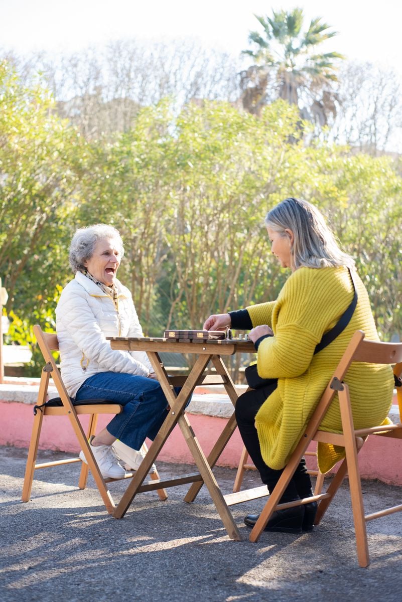 Two senior women laughing together while playing dominoes outdoors, representative image