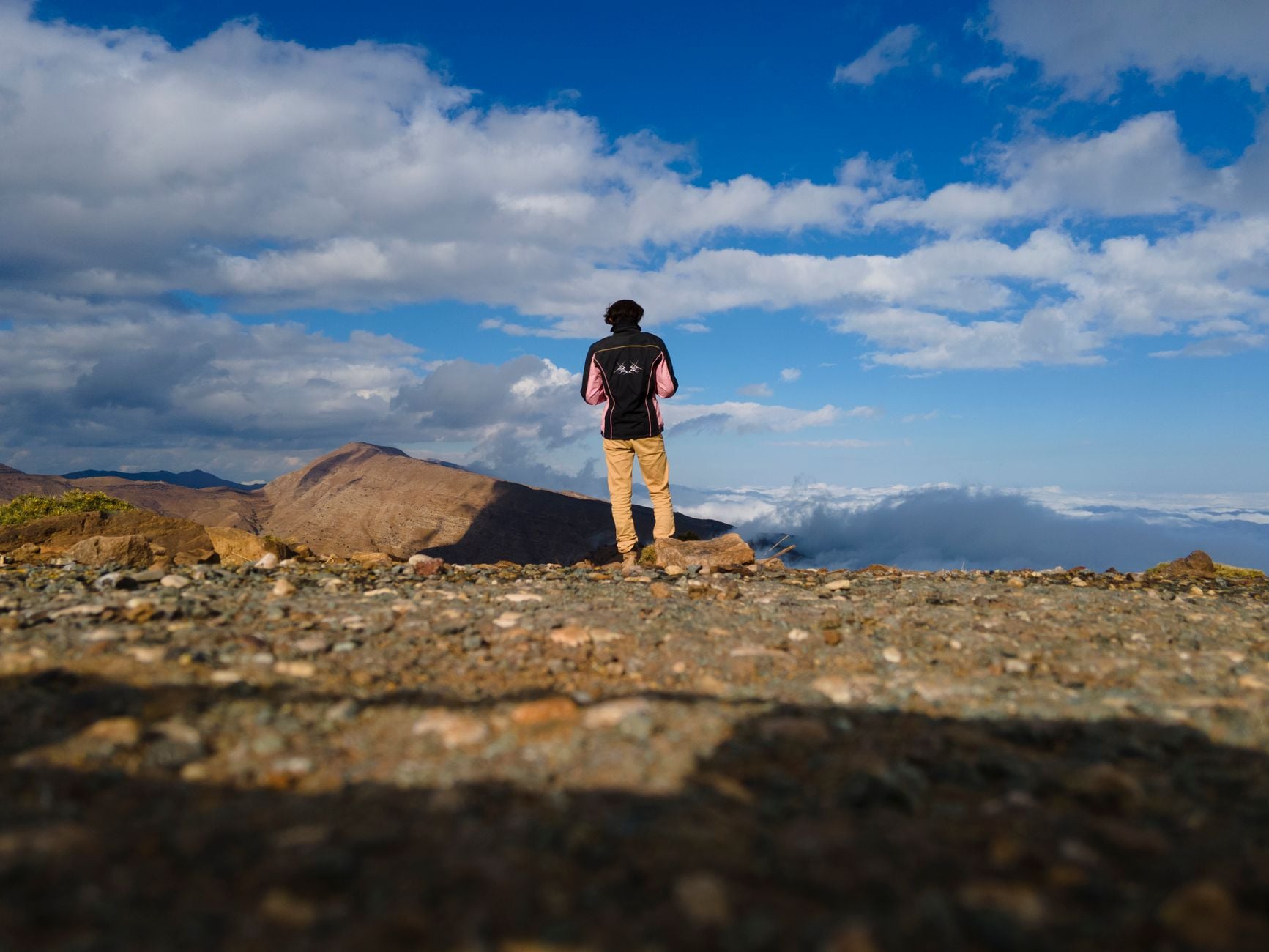 A lone hiker stands at a mountain summit looking out over clouds and distant peaks.