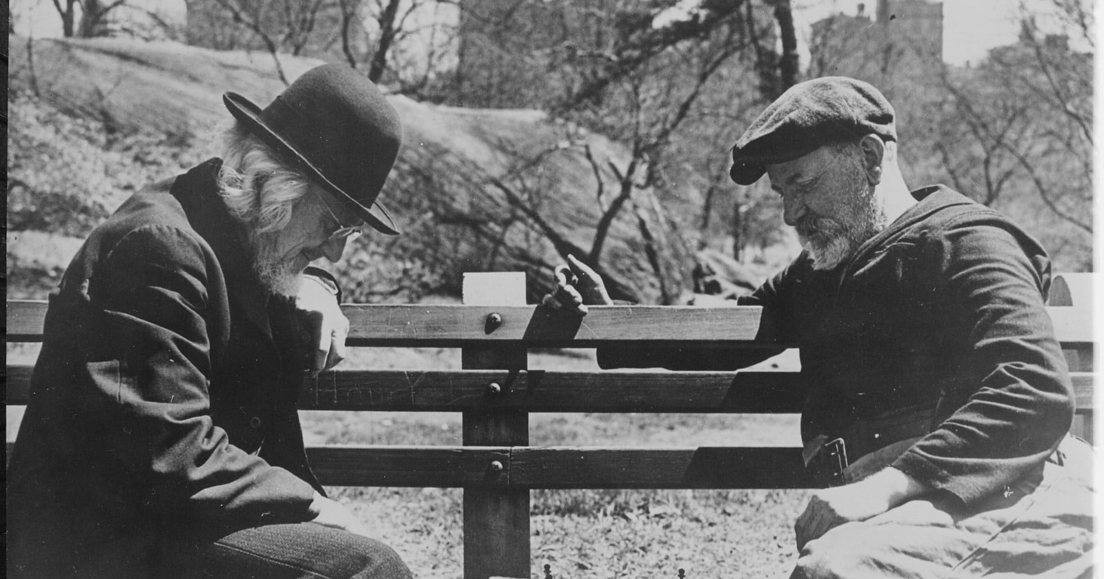 Two older men in coats and caps lean toward each other over a chess game on a park bench.