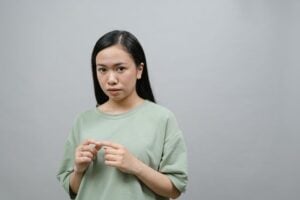 A pensive young woman stands in a studio with a neutral gray background, expressing contemplation