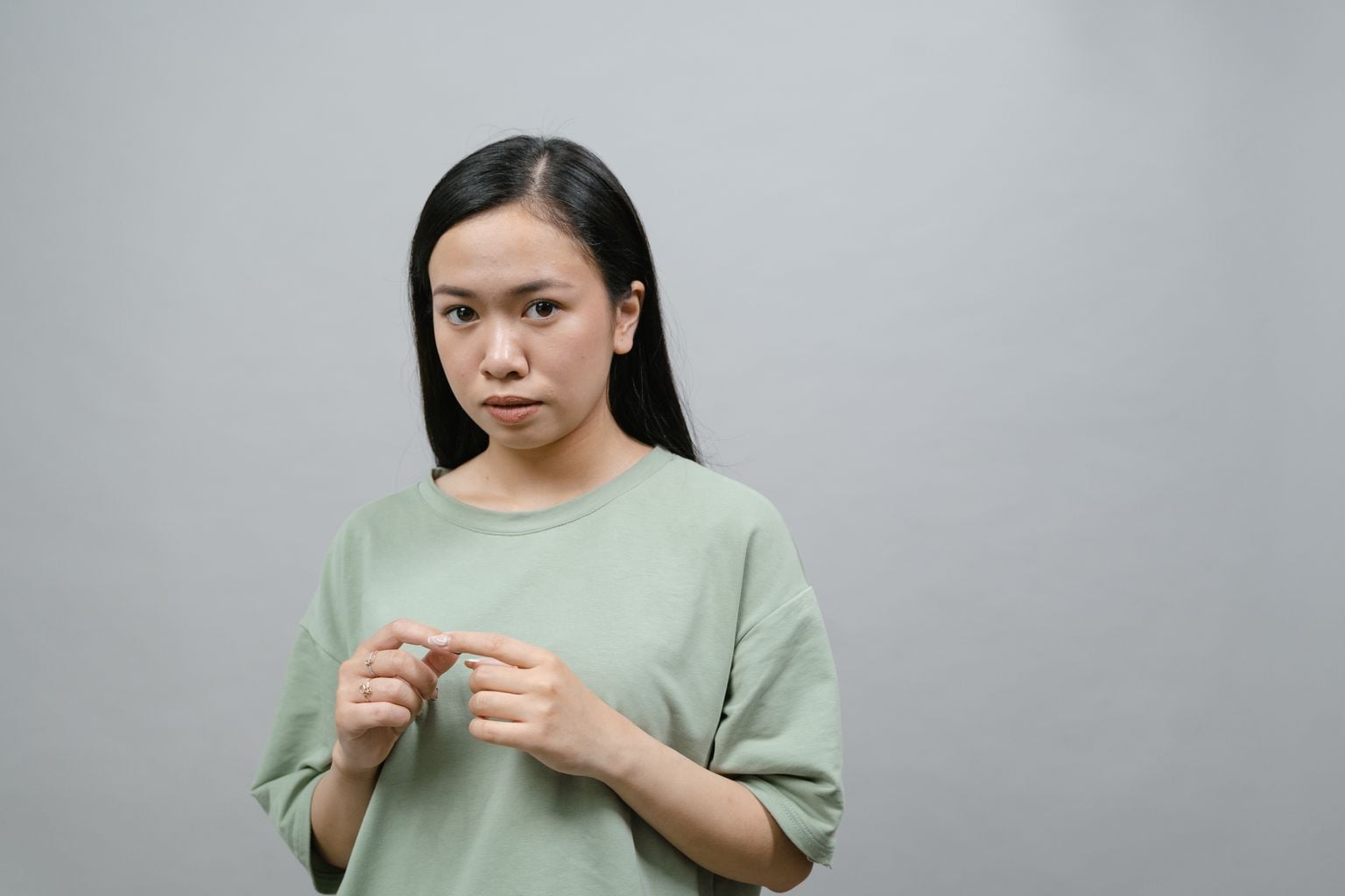 A pensive young woman stands in a studio with a neutral gray background, expressing contemplation